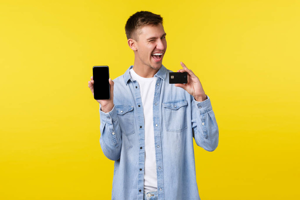 Technológia, életmód és reklám koncepció. Pimasz jóképű férfi, hogy online rendelés, vásárlás az interneten a mobil app, mutatja okostelefon képernyő és hitelkártya, sárga háttér - Fotó, kép