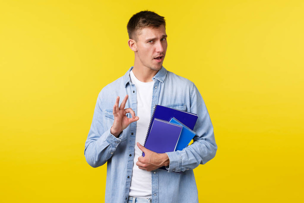 Education, courses and university concept. Handsome assured blond guy encourage enroll this college, showing okay gesture as guarantee you will like it, holding notebooks, yellow background - Фото, зображення