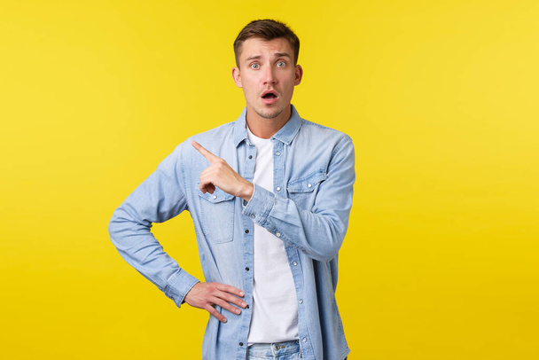 Worried and concerned blond handsome guy looking insecure and anxious, pointing finger upper left corner stare camera alarmed, feeling scared as standing over yellow background - Photo, Image