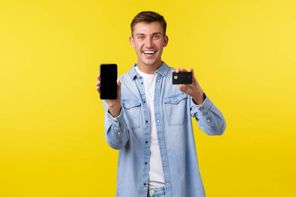 Tecnología, estilo de vida y concepto de publicidad. Sonriente cliente masculino guapo, estudiante mostrando aplicación super cool, pantalla del teléfono inteligente y tarjeta de crédito, recomendando tienda, fondo amarillo
 - Foto, Imagen