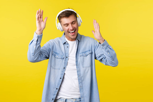 Lifestyle, summer holidays, technology concept. Excited happy handsome guy in casual clothes, listening music in wireless headphones, dancing and smiling, enjoying favorite song, yellow background - Photo, Image