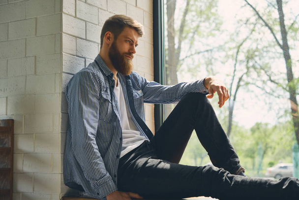 Young attractive bearded man rests and looks out the window. Enjoy the morning afternoon. Healthy lifestyle concept. - Fotó, kép