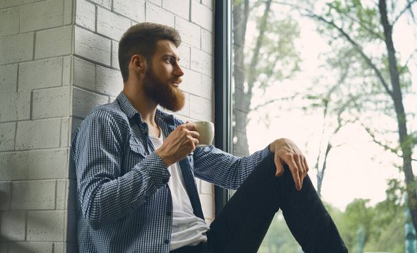 若い魅力的な髭の男が残り、窓の外を見ています。朝の午後をお楽しみください。健康的なライフスタイルのコンセプト。距離を調べる新鮮なブレースコーヒーを飲む - 写真・画像