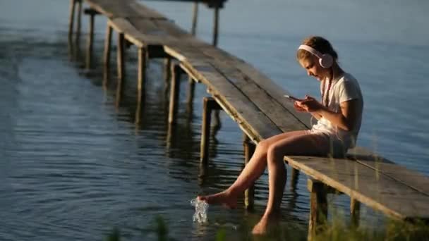 Ένα μικρό κορίτσι που φοράει ακουστικά με ένα smartphone κάθεται σε μια ξύλινη προβλήτα στη λίμνη κατά τη διάρκεια του ηλιοβασιλέματος. Εξωτερική αναψυχή το καλοκαίρι. Η έννοια της ευτυχισμένης παιδικής ηλικίας. - Πλάνα, βίντεο