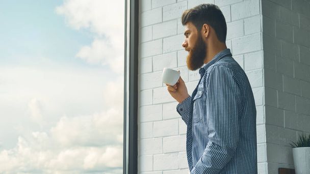 Casual young bearded man drinking coffee at work Coffee really gives more energy. Bearded man enjoy coffee in morning looking at the window. Happy hipster hold cup of hot energy drink. - 写真・画像