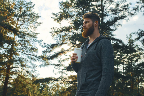 Bearded hipster guy wearing casual clothes enjoys relaxing on a summer weekend. Young bearded man holding coffee to take away at early morning in sunny park, sunlight, blurred background - Foto, imagen