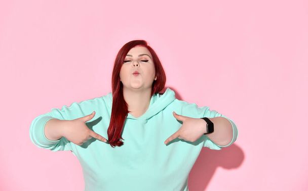 Fatty young lady in stylish blue sweater pointing on herself with both index fingers showing a kiss by her lips. Waist-up shot isolated on pink - Photo, Image