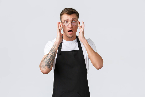 Small retail business owners, cafe and restaurant employees concept. Worried and confused cute barista, waiter put on glasses, open mouth and looking alarmed, having trouble, problem at work - Photo, Image