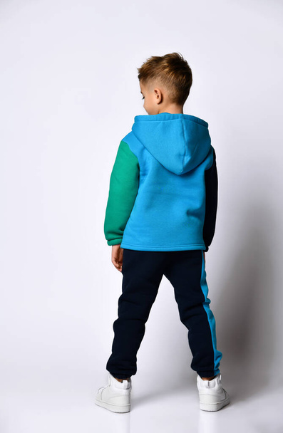 Little boy in colorful sport suit, sneakers. He looking back, posing standing back to camera, isolated on white studio - Φωτογραφία, εικόνα