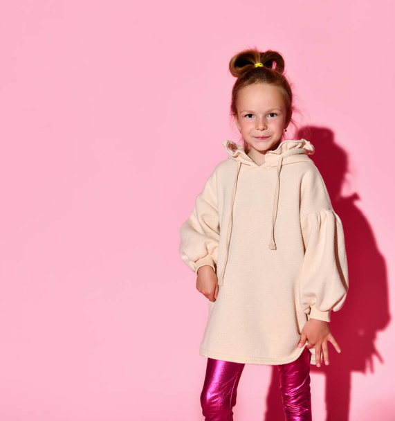 Cute little girl with updo posing with confident look in pale-pink sweatshirt and glossy leggings. Three quarter length isolated on pink, copy space - Fotografie, Obrázek