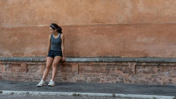 jong tiener meisje met hoofdband, shorts, tanktop en sneakers zitten op baksteen bank in Bolgona Italië - Foto, afbeelding
