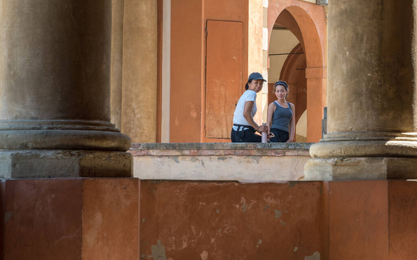 Mujer asiática sentada en la pared con hija adolescente de pie junto a ella rodeada por un edificio en Bolonia, Italia
 - Foto, Imagen