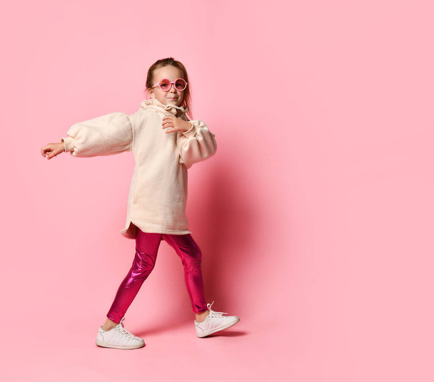 Adorable little girl wearing glasses, warm pale-pink hoodie and leggings is marching with a light smile. Full length shot isolated on pink, copy space - 写真・画像