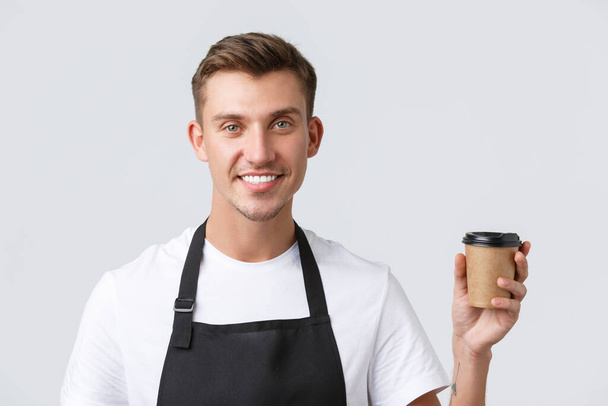 Cafe and restaurants, coffee shop owners and retail concept. Close-up shot of friendly handsome barista, waiter serving takeaway drink to client, holding paper cup and smiling, white background - Foto, imagen