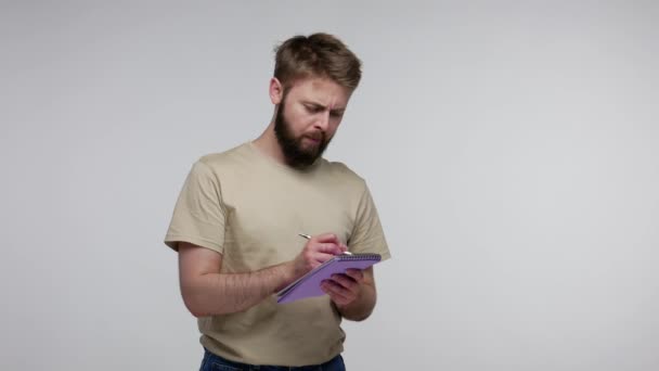 Bearded man in t-shirt thinking, remembering and taking notes in paper notebook, writing creative idea, smiling inspired happy, making to-do list plans. indoor studio shot isolated on gray background - Séquence, vidéo
