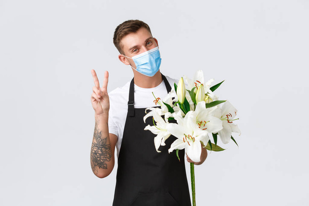Coronavírus, negócio de distanciamento social durante o conceito de pandemia covid-19. Bonito e amigável florista, vendedor em show de flores, mostrando sinal de paz sorrindo e segurando lírios buquê, use máscara médica
 - Foto, Imagem