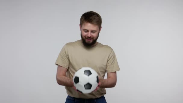 Crazy delighted football fan bearded guy in t-shirt holding soccer ball and shouting for joy, excited about winning goal, cheering for favorite sport game. studio shot isolated on gray background - Video
