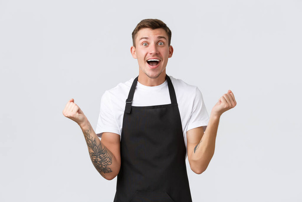 Employees, grocery stores and coffee shop concept. Happy and relieved smiling handsome barista, waiter in black apron, fist pump in rejoice, triumphing victory or great positive news - Photo, Image