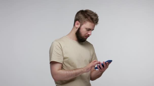 Mobile user, concentrated attentive bearded guy typing message using cellphone to communicate online, chat in social network, scrolling media pictures and smiling. studio shot isolated gray background - 映像、動画