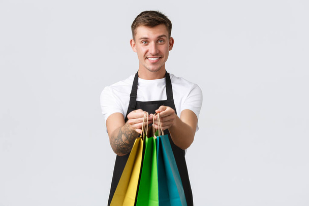 Retail store, shopping and employees concept. Friendly handsome and polite salesman handing over eco-bags with purchased items to client in shop, standing white background - Photo, Image