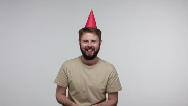 Optimistic joyful bearded guy with funny cone hat laughing and blowing confetti glitters, celebrating birthday congratulating on anniversary, festive mood. studio shot isolated on gray background - Materiaali, video