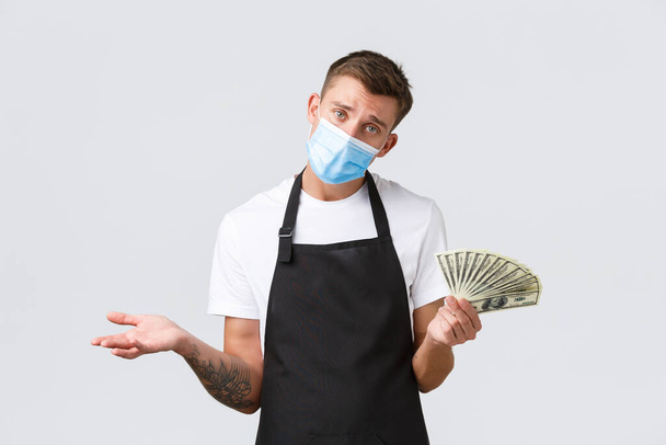 Small retail business, employees and coffee shop concept. Upset and gloomy barista, cafe owner or waiter in medical mask counting his money, shrugging with hands in cash, white background - Photo, Image