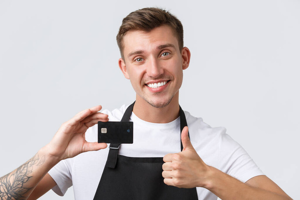 Cafe and restaurants, coffee shop owners and retail concept. Cheerful handsome salesman, store clerk in black apron showing thumbs-up in approval, recommend paying for credit card - Foto, Imagem