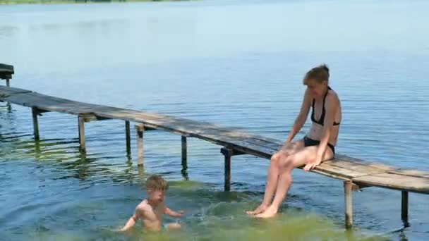 Egy kisfiú vizet fröcsköl az anyjára a tónál. Szabadtéri kikapcsolódás nyáron. A boldog gyermekkor fogalma. - Felvétel, videó