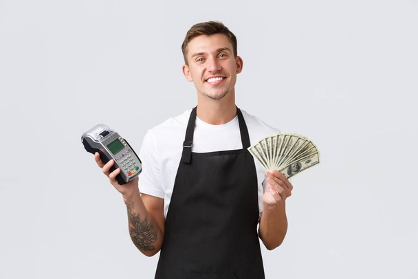 Small retail business, payments and employees concept. Friendly handsome salesman, barista or waiter in black apron giving client POS terminal as customer paying, holding money - Photo, Image