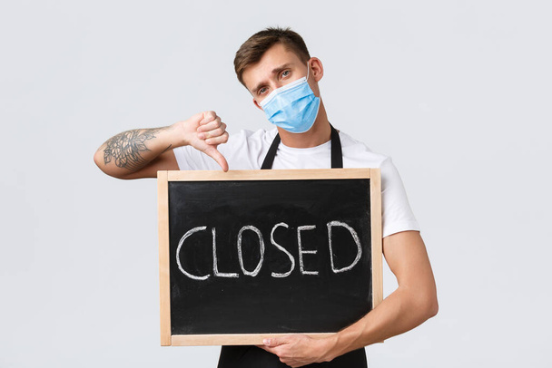 Small retail business owners, covid-19 and social distancing concept. Disappointed sad barista, waiter in medical mask showing thumbs-down as inform clients restaurant closed, white background - Foto, imagen