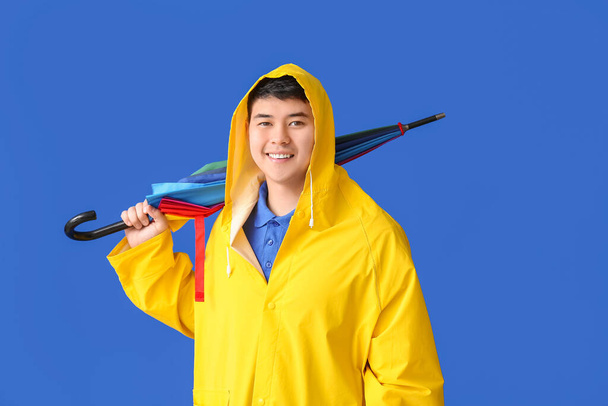 Junger asiatischer Mann im Regenmantel und mit Regenschirm auf farbigem Hintergrund - Foto, Bild