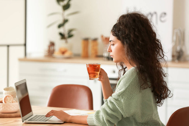 Mooie jonge vrouw drinken hete thee tijdens het werken op laptop thuis - Foto, afbeelding