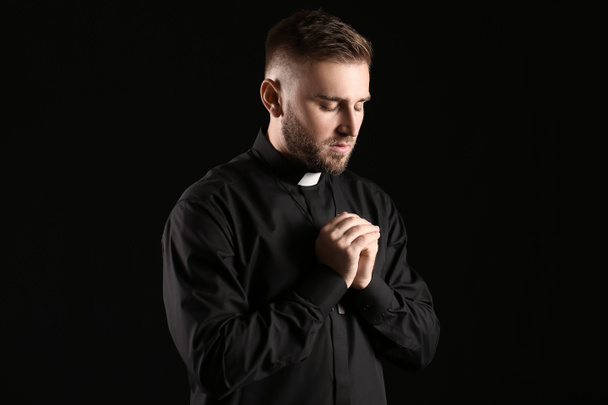 Young priest praying to God on dark background - Фото, изображение