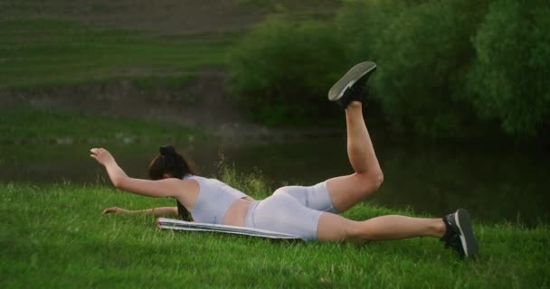 Το πρωί στο πάρκο, μια γυναίκα με αθλητικά ρούχα ξαπλωμένη μπρούμυτα σηκώνει τα χέρια και τα πόδια της με τη σειρά. Ασκήσεις για ένα όμορφο σώμα. Σταθμοί μυών - Πλάνα, βίντεο