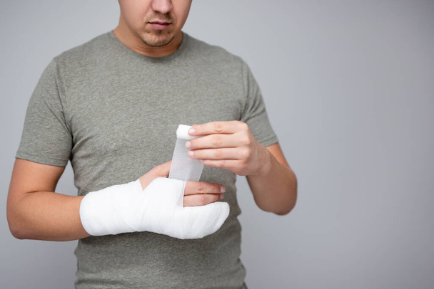 fracture and first aid concept - close up of young man bandaging his hand after accident and copy space over gray wall background - Photo, Image