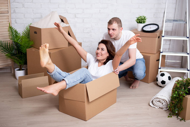 moving day and happiness concept - happy couple having fun and riding in cardboard boxes in their new home or flat - Photo, image