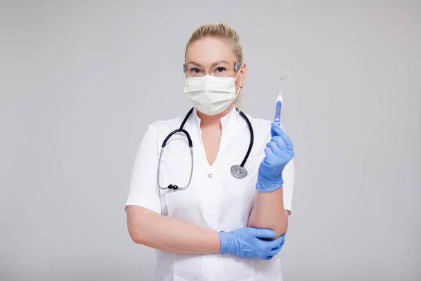 Концепція охорони здоров'я та пандемії коронавірусу - жінка-лікар або медсестра в білій формі та захисній масці, що тримає термометр на сірому фоні з копіювальним простором
 - Фото, зображення
