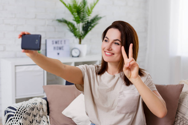 πορτρέτο της νεαρής γυναίκας που δείχνει το σήμα της νίκης και τη λήψη selfie φωτογραφία με έξυπνο τηλέφωνο στο άνετο σαλόνι στο σπίτι - Φωτογραφία, εικόνα