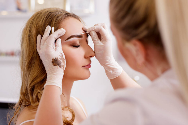 Make-up und Beauty-Konzept - junge schöne blonde Frau bekommt Augenbrauen-Malverfahren im Schönheitssalon - Foto, Bild