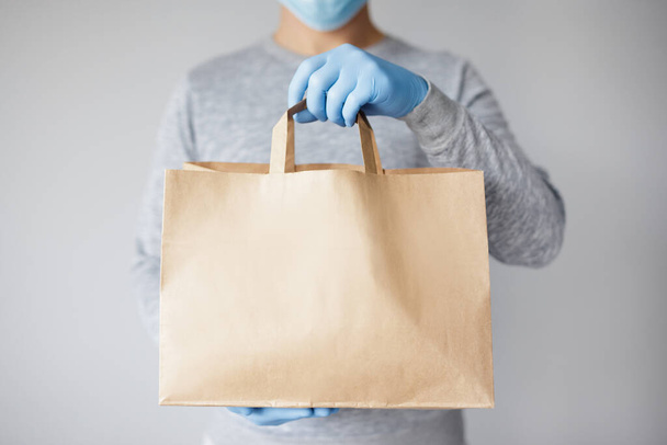 coronavirus concept - courier or food delivery man in medical mask and gloves holding paper package over white background - Photo, Image