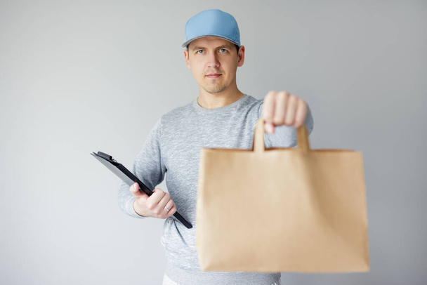 delivery concept - portrait of young delivery man holding clipboard and giving paper bag over white background - Photo, Image