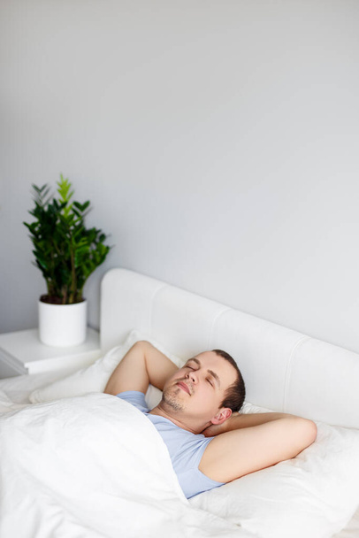 νεαρός όμορφος άντρας κοιμάται στο κρεβάτι στο σπίτι και να αντιγράψετε χώρο πάνω από λευκό τοίχο - Φωτογραφία, εικόνα