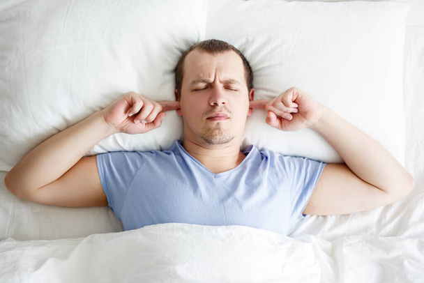 Gürültücü komşular ve baş ağrısı - uykucu yorgun genç adam yatakta uzanıyor ve parmaklarıyla kulaklarını kapatıyor gürültüden korunuyor - Fotoğraf, Görsel