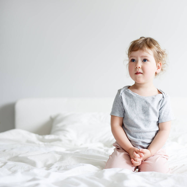 мила маленька дівчинка сидить на ліжку і мріє або думає про щось, копіює простір на білому тлі стіни
 - Фото, зображення