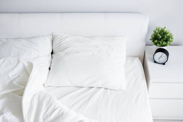 widok z góry puste łóżko niepościelone z białym kocem i dwie poduszki, szafka nocna z budzikiem i garnek roślin domowych w domu lub hotelu - Zdjęcie, obraz