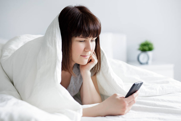 portrait de jeune belle femme utilisant téléphone intelligent couché dans le lit à la maison - Photo, image