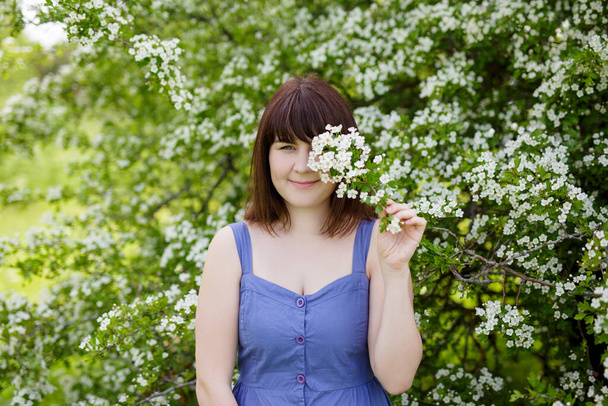 portrait of young beautiful woman covering eye with blooming tree branch in summer garden - Photo, Image