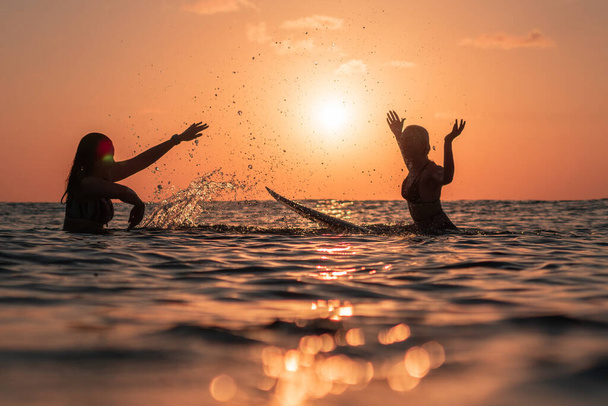 Portrait from the water of two surfer girls with beautiful bodies on surfboards splashe water in the ocean at colourful sunset time in Bali - Valokuva, kuva