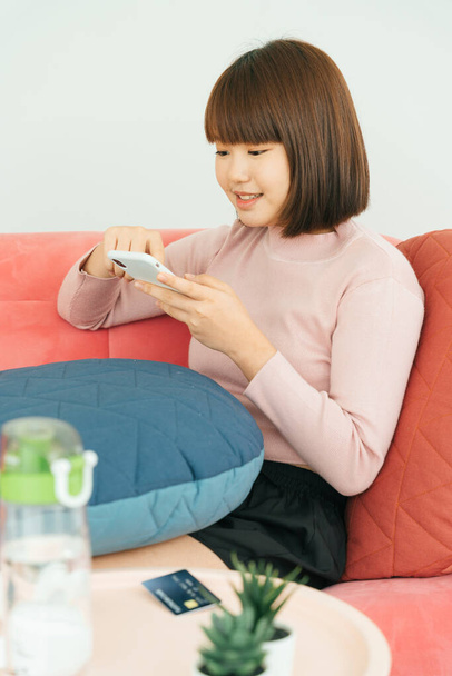 Hermosa joven asiático tailandés negro pelo corto mujer usando jugando teléfono inteligente sentado en un sofá en la sala de estar
. - Foto, Imagen