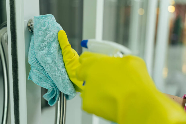 Schoonmaakster draagt gele beschermende handschoenen bij het schoonmaken van de deur ingang met vod en wasmiddel spray fles. Covid-19 Coronaviruspreventie. - Foto, afbeelding